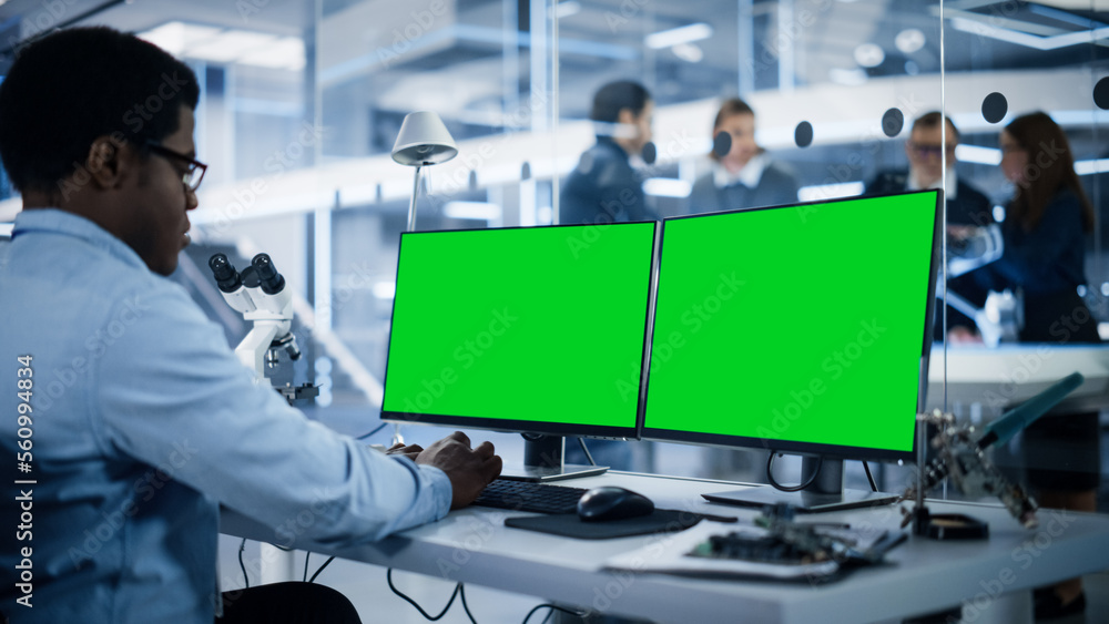 Software Developer Working on Computer with Green Screen Mock Up Display. Scientific Lab, Engineering Research Center with Specialists Working on Mobile Robot Development in the Background. Software Developer Working on Computer with Green Screen Mock Up Display. Scientific Lab, Engineering Research Center with Specialists Working on Mobile Robot Development in the Background.