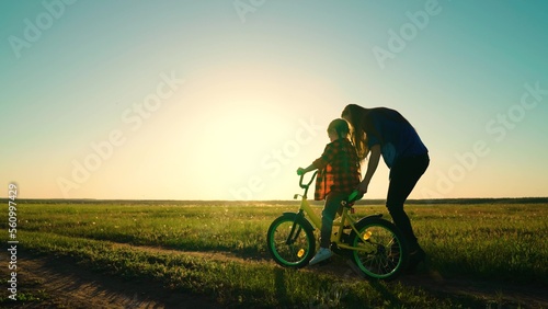 Active Mom teaches her little daughter to ride bike, sunset. Mother teaches her child to keep balance while sitting on bicycle. Childhood dream of riding bike. Family life, mom, baby, parental support