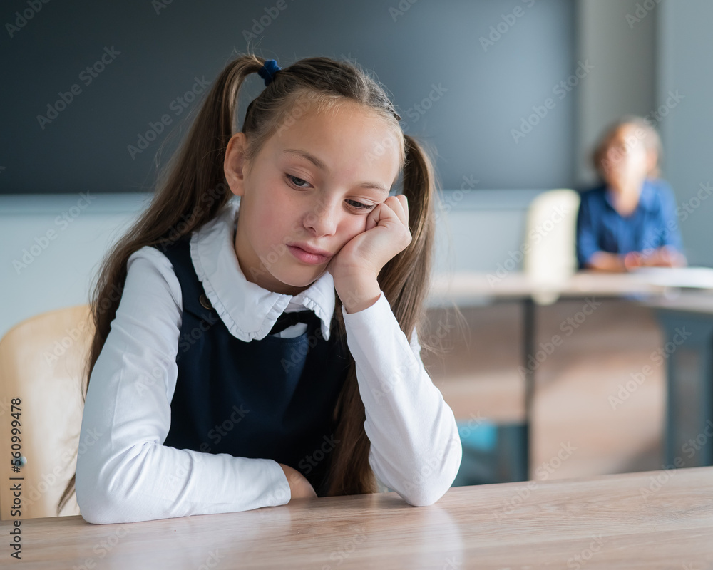 Little caucasian girl is bored at the lesson at school. The schoolgirl is sitting at her desk and the teacher is sitting in the background. Little caucasian girl is bored at the lesson at school. The schoolgirl is sitting at her desk and the teacher is sitting in the background.