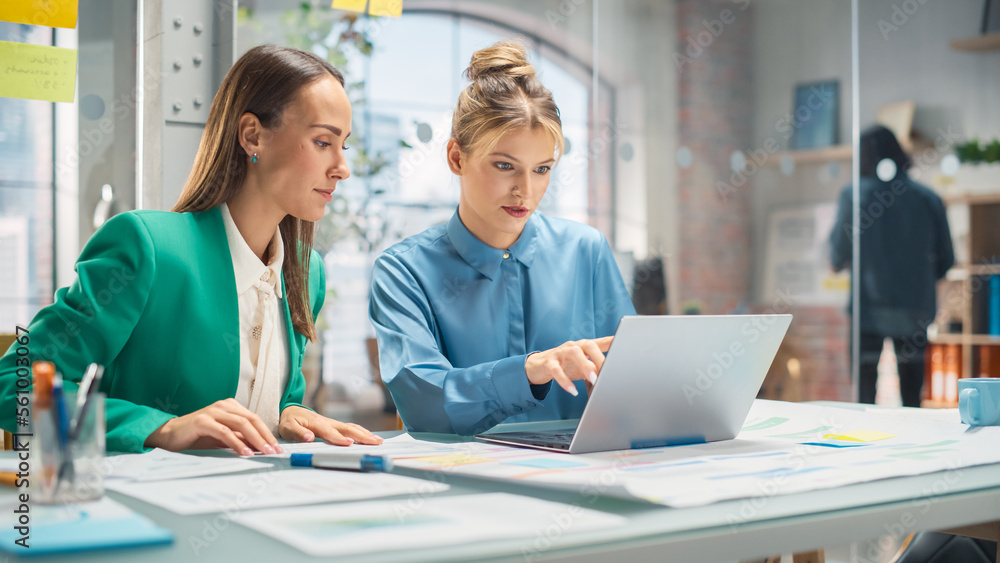 Two White Females Have a Discussion in Meeting Room Behind Glass Walls in an Agency. Creative Director and Project Manager Compare Business Results and App Designs on Laptop in an Office Two White Females Have a Discussion in Meeting Room Behind Glass Walls in an Agency. Creative Director and Project Manager Compare Business Results and App Designs on Laptop in an Office
