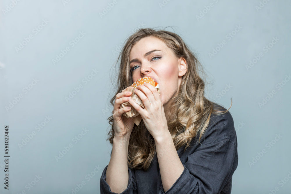 A young woman eats a hamburger with appetite. fast junk delicious food. Grey background. Close-up. A young woman eats a hamburger with appetite. fast junk delicious food. Grey background. Close-up.