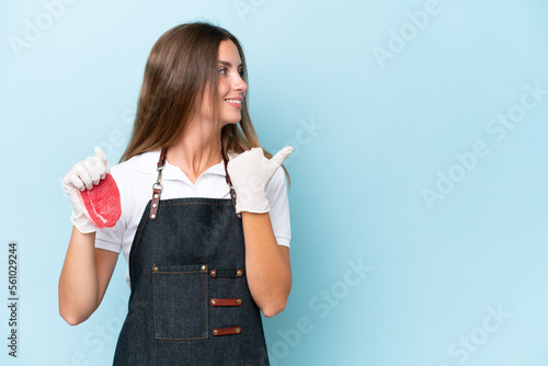 Butcher caucasian woman wearing an apron and serving fresh cut meat isolated on blue background pointing to the side to present a product