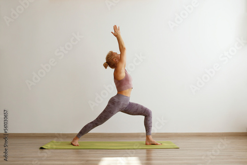 Sporty adult woman practicing hatha yoga at home. Fit middle aged yogini doing the warrior I pose. White wall background, copy space, close up.