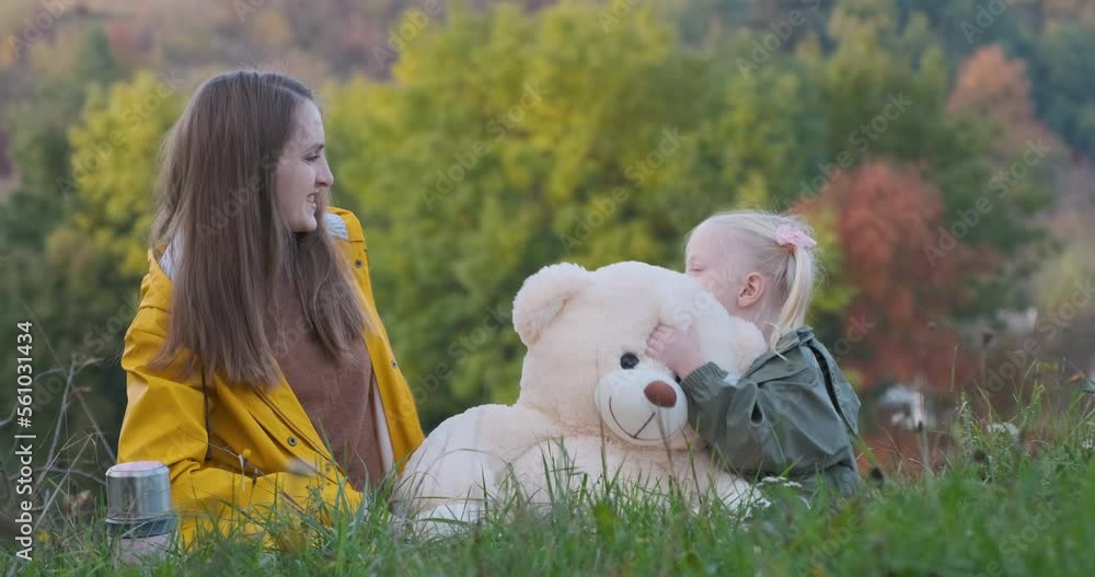 Young woman laughs and plays with little daughter sitting in grass in nature. Blonde girl plays large teddy bear outside Young woman laughs and plays with little daughter sitting in grass in nature. Blonde girl plays large teddy bear outside