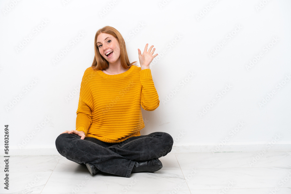 Young redhead woman sitting on the floor isolated on white background saluting with hand with happy expression