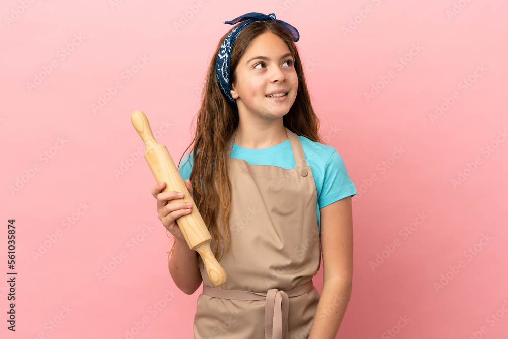 Little caucasian girl holding a rolling pin isolated on pink background thinking an idea while looking up Little caucasian girl holding a rolling pin isolated on pink background thinking an idea while looking up