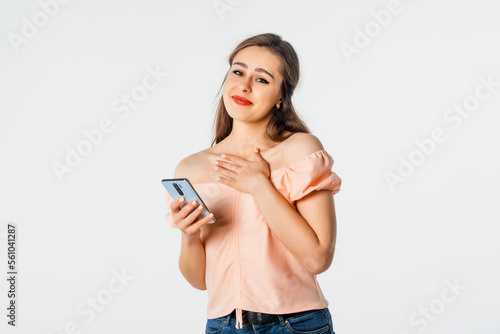 Joyful beautiful woman holding cellphone and gesturing hand while standing over white background. Pretty girl holding cell phone and smiling