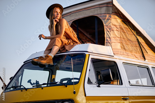 Dreamy calm female with long hair sits on roof of yellow van for trip, enjoy, having rest alone, in countryside in rural nature. holidays at summer, sunset. tourism, travel, people lifestyle concept photo