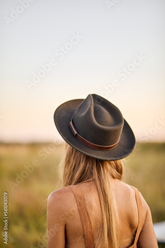 Attractive long-haired woman in dress and hat having rest enjoying trip at summer field, rear view on caucasian slender lady during travel, take easy the life, enjoy and relax summertime.