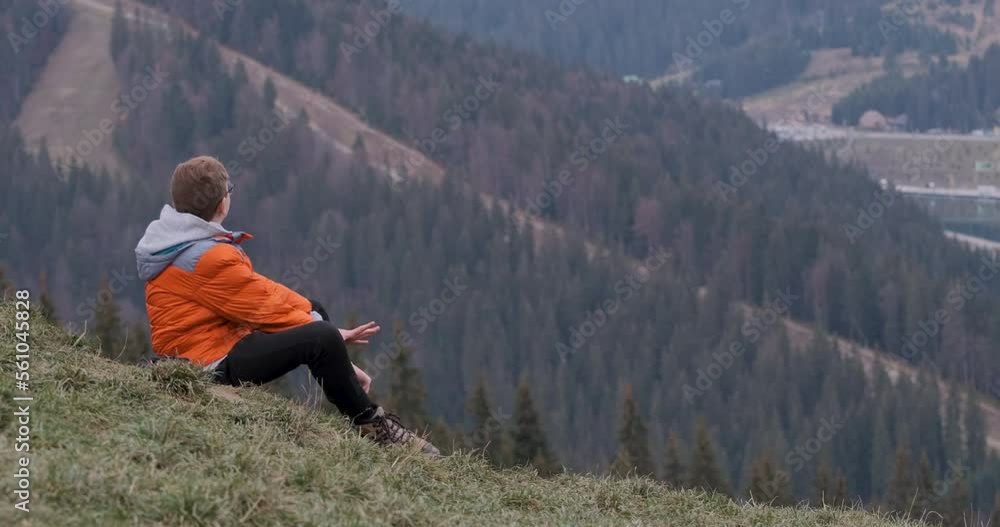 Teenage boy in glasses and bright orange jacket rests on grass on mountainside. Child in unity with nature