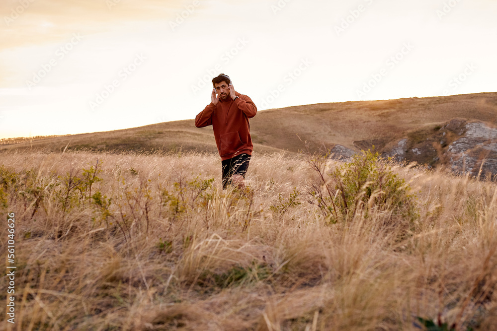 man walking alone in field listening to music, at summer or autumn season, handsome brunette guy in red casual hoodie having rest in countryside, healthy and fit male thinking, in contemplation