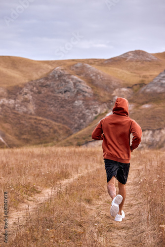Rear view on athlete adult man in red hoodie is running jogging, listen to music, enjoy sport. wonderful landscape, mountains in the background. man is in action, strong and fit. cardio training © Roman