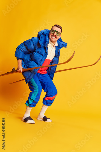 Cheerfully smiling. Portrait of handsome man in blue winter jacket posing with skis over bright yellow background. Concept of leisure time, winter sport, hobby