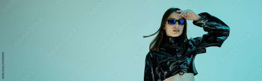 stylish young woman in black latex clothing and sunglasses standing isolated on blue, banner.