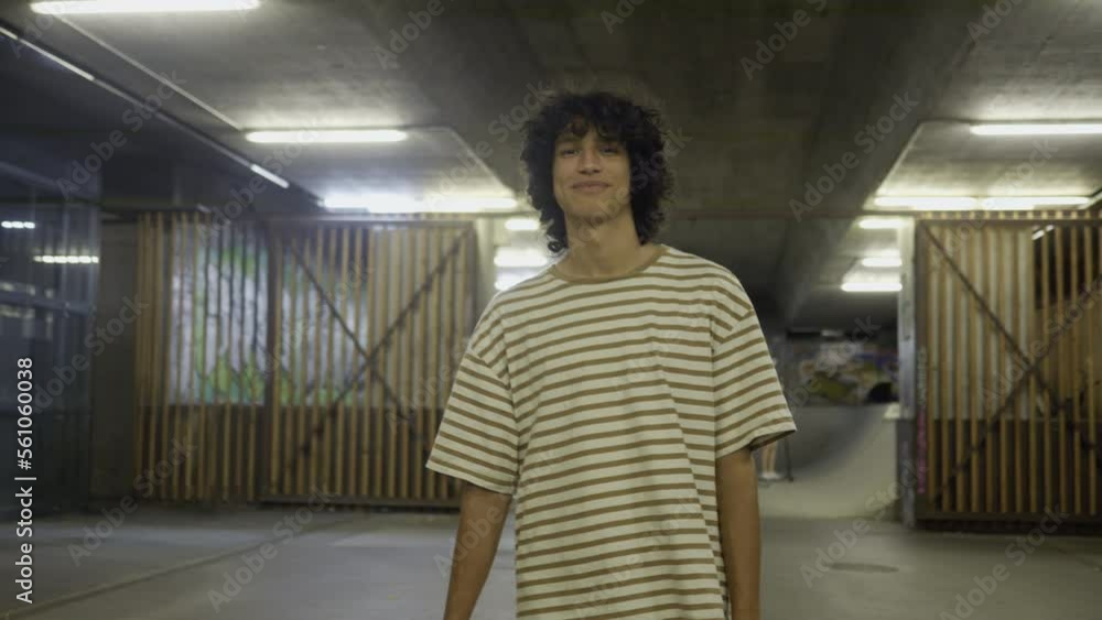Smiling boy picking up skateboard and smiling in indoor skatepark portrait, handheld tilt up shot. Cheerful child standing, flipping skate and looking at camera, front view