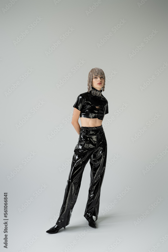 Obraz premium Full length of stylish woman in jewelry headwear and latex clothes standing on grey background.