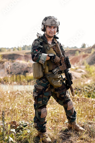 equipped camouflaged soldier with rifle gun standing and posing outdoors, alone. portrait of young male with weapon looking at camera confidently. military forces, defense concept