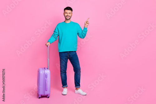 Full length photo of young positive handsome man wear stylish jumper with luggage direct finger mockup trip agency isolated on pink color background