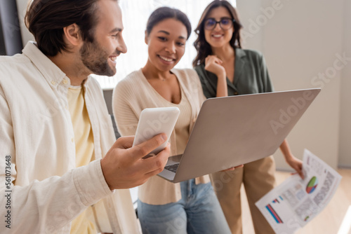 smiling manager holding smartphone near interracial businesswomen with laptop and documents on blurred background.