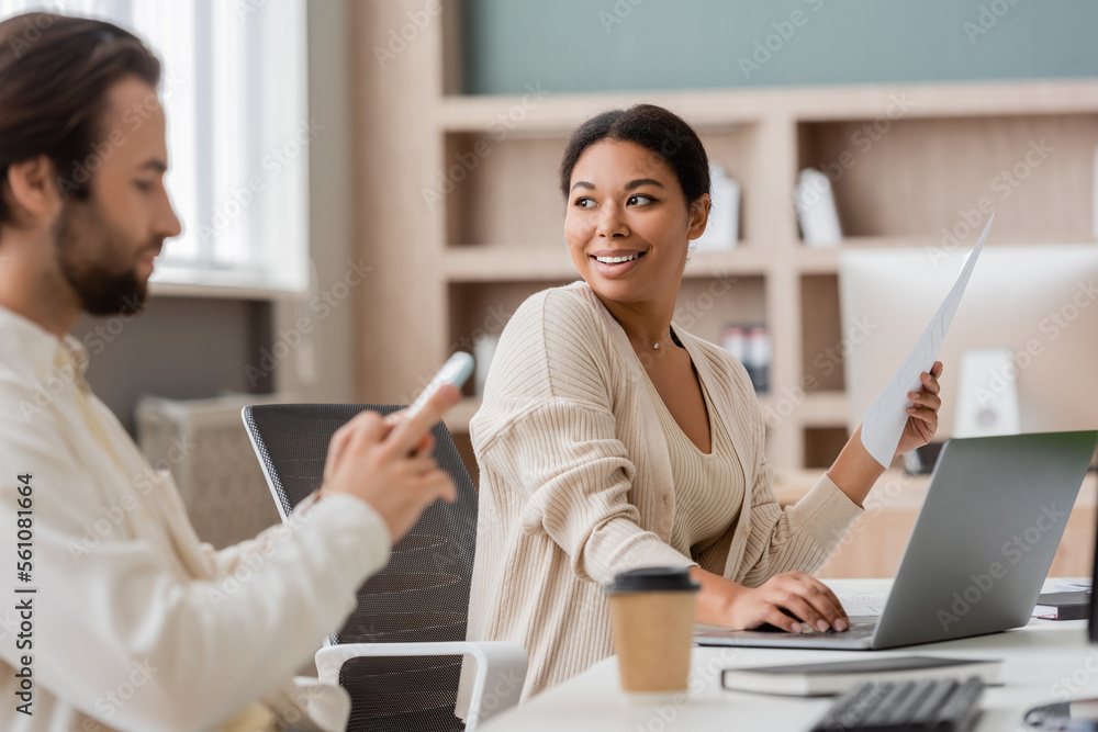 smiling multiracial businesswoman with document looking away near laptop and blurred colleague in office. smiling multiracial businesswoman with document looking away near laptop and blurred colleague in office.