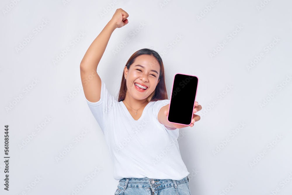 Naklejka premium Beautiful excited Asian girl wearing casual white t-shirt showing mobile phone with blank white screen, raising fist, celebrating good luck isolated on white background. People lifestyle concept