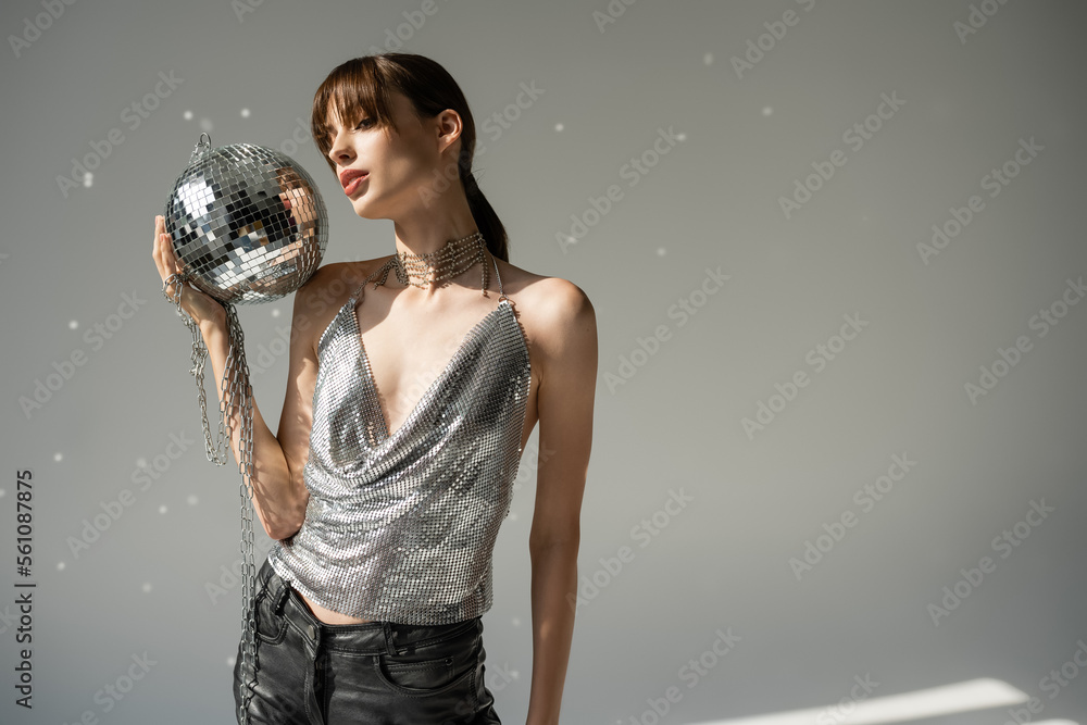 Naklejka premium young woman in trendy top and leather pants holding chain with disco ball on grey.