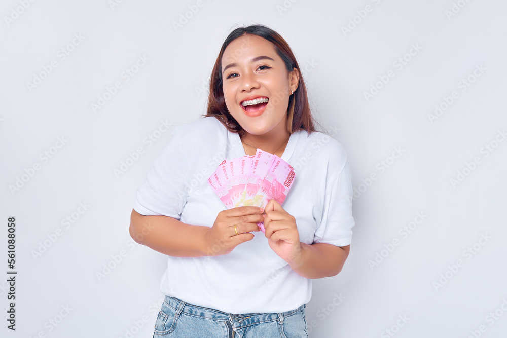 Excited young blonde woman girl Asian wearing casual white t-shirt holding money rupiah banknotes and smiling at the camera isolated on white background. Profit and wealth concept Excited young blonde woman girl Asian wearing casual white t-shirt holding money rupiah banknotes and smiling at the camera isolated on white background. Profit and wealth concept