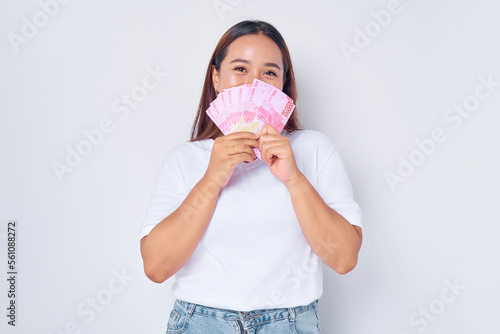 Smiling young blonde woman girl Asian wearing casual white t-shirt covering her face with money rupiah banknotes isolated on white background. people lifestyle concept