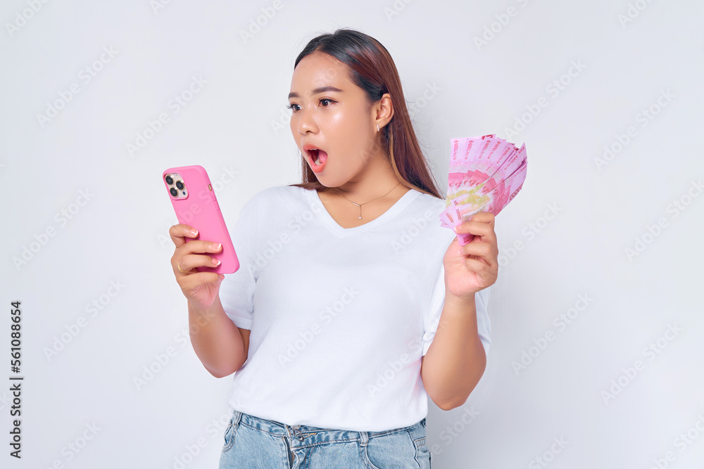 Shocked young blonde woman girl Asian wearing casual white t-shirt using mobile phone and holding money rupiah banknotes isolated on white background. Fast credits concept