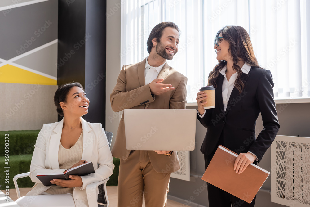 smiling manager with laptop talking to colleague standing with coffee to go and folder near multiracial businesswoman in office. smiling manager with laptop talking to colleague standing with coffee to go and folder near multiracial businesswoman in office.