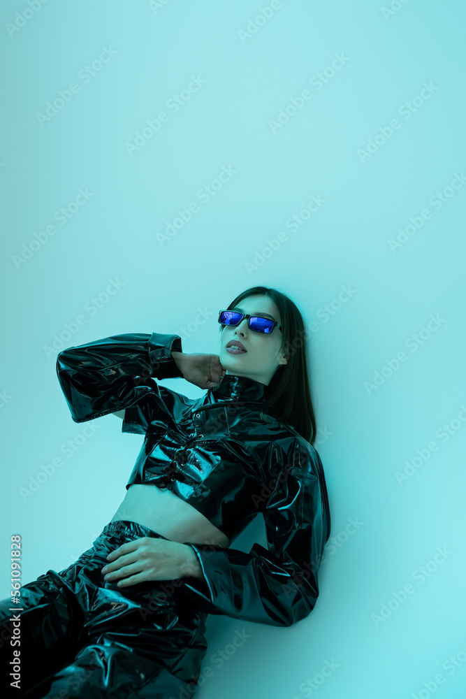 stylish young woman in sunglasses and black latex clothing sitting on blue background. stylish young woman in sunglasses and black latex clothing sitting on blue background.