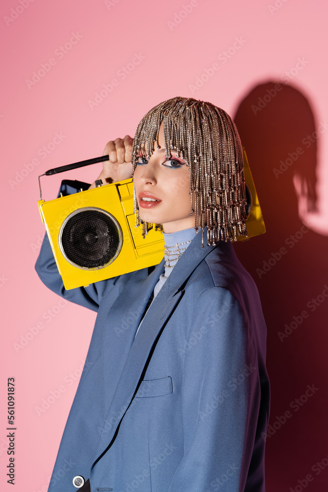 Obraz premium Trendy woman in jacket and jewelry headwear holding boombox on pink background.
