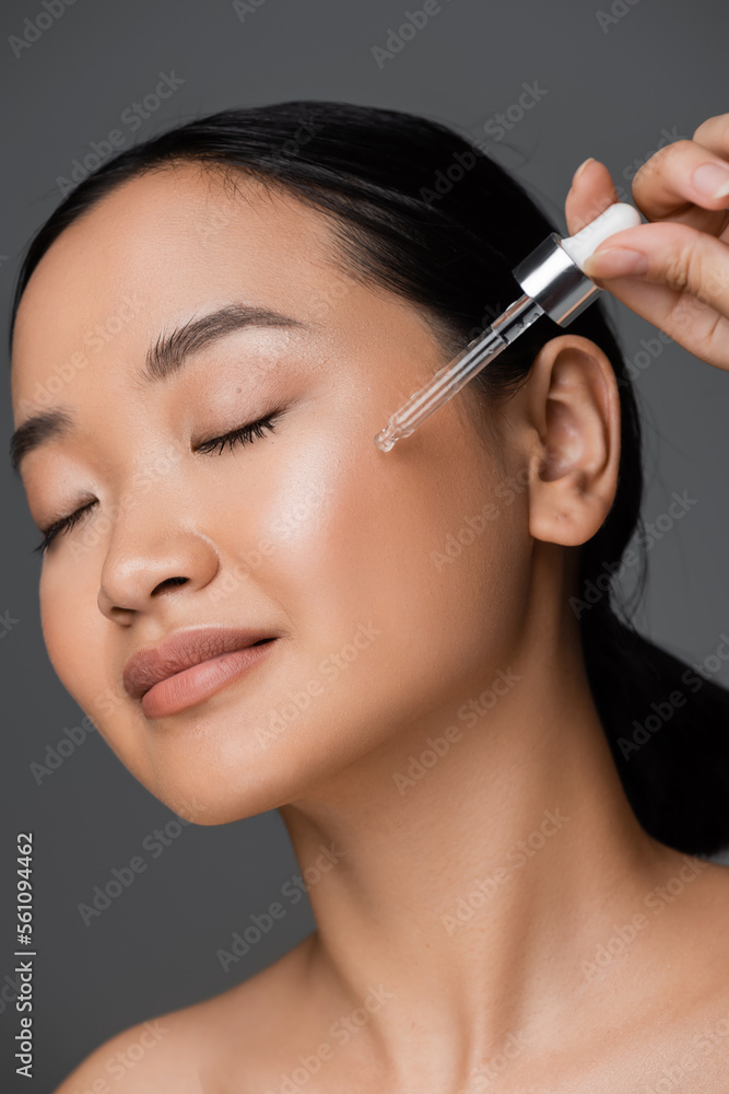 young asian woman with closed eyes applying cosmetic serum on clean face isolated on grey.