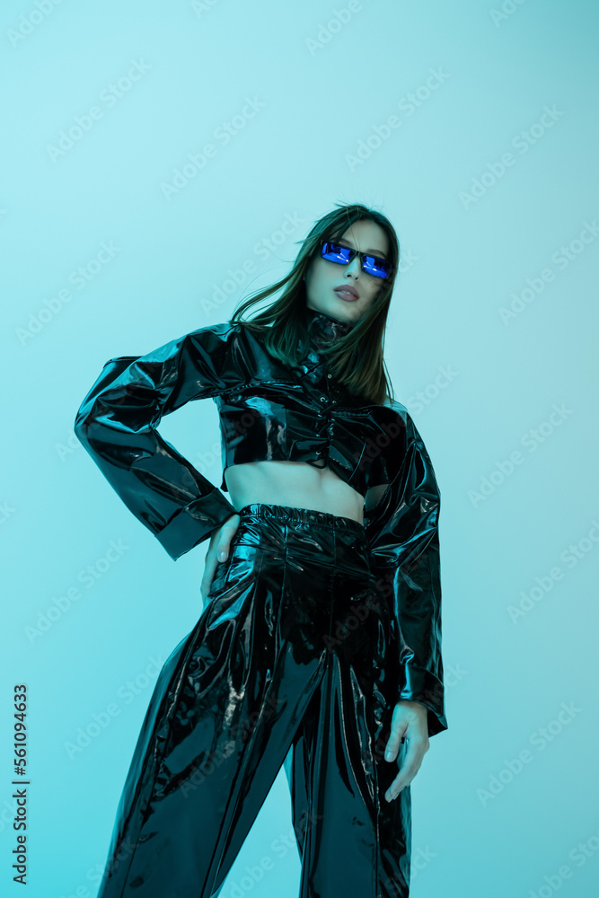 low angle view of stylish young woman in latex clothing posing with hand on hip isolated on blue.