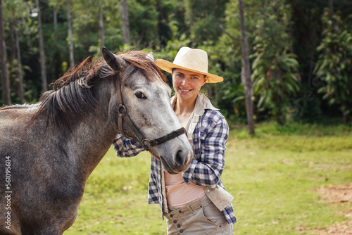 Young woman in shirt and straw hat posing next to gray horse, jungle trees background - horseriding, ranch at Isalo Park, Madagascar