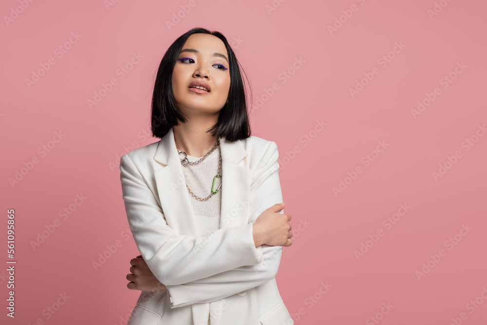 brunette asian woman in white jacket and necklaces standing with crossed arms and looking away isolated on pink. brunette asian woman in white jacket and necklaces standing with crossed arms and looking away isolated on pink.