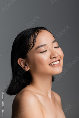 young asian woman with bare shoulders and nude makeup smiling with closed eyes isolated on grey.