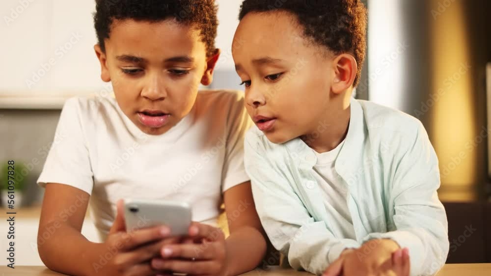 Adorable cute african american little boys brothers hold smartphone scrolling social media use educational children app application having fun with mobile phone gadget or play game together at home Adorable cute african american little boys brothers hold smartphone scrolling social media use educational children app application having fun with mobile phone gadget or play game together at home