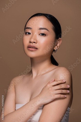 Young asian woman in top touching shoulder and looking at camera isolated on brown.