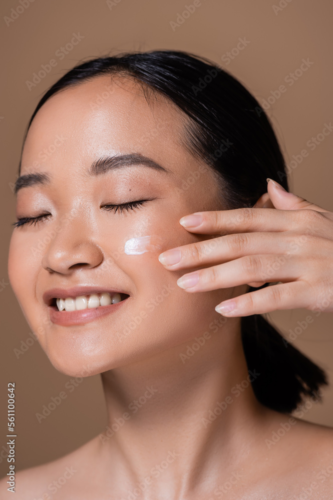 Smiling asian woman applying face cream isolated on brown.