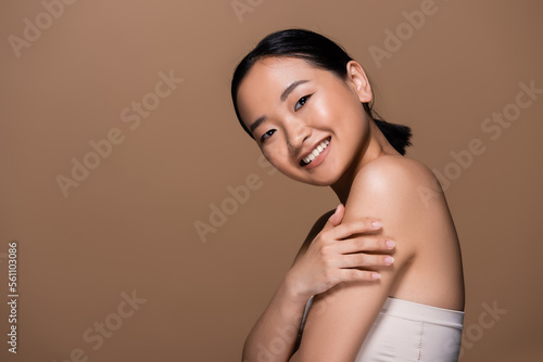 Positive asian woman in top touching arm and looking at camera isolated on brown.