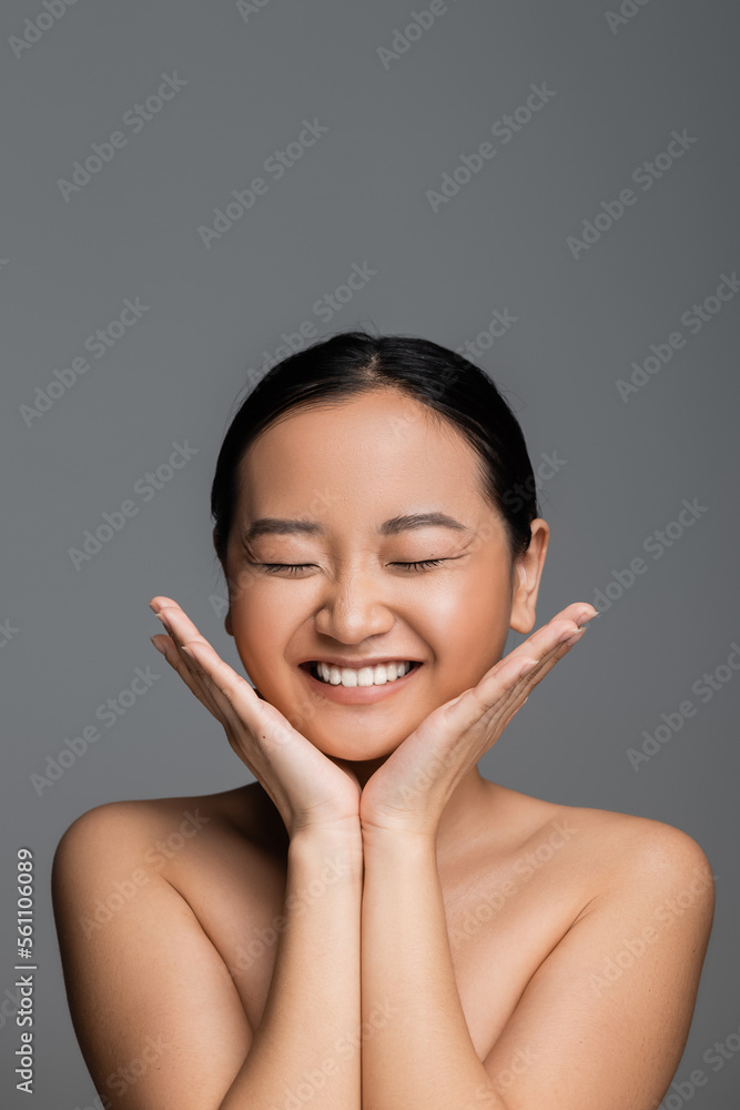 excited asian woman with closed eyes and nude makeup holding hands near face isolated on grey. excited asian woman with closed eyes and nude makeup holding hands near face isolated on grey.