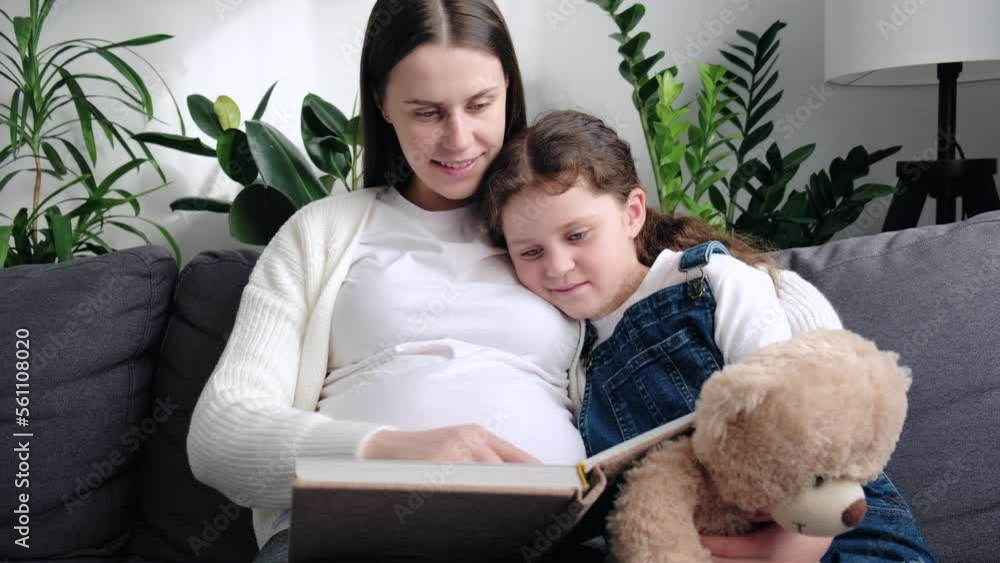 Happy caring mom and daughter engaged in fairytale, learning story, reading book together sitting on cozy sofa. Pregnant mom teaching girl child to read, enjoying home activity. Entertainment concept Happy caring mom and daughter engaged in fairytale, learning story, reading book together sitting on cozy sofa. Pregnant mom teaching girl child to read, enjoying home activity. Entertainment concept