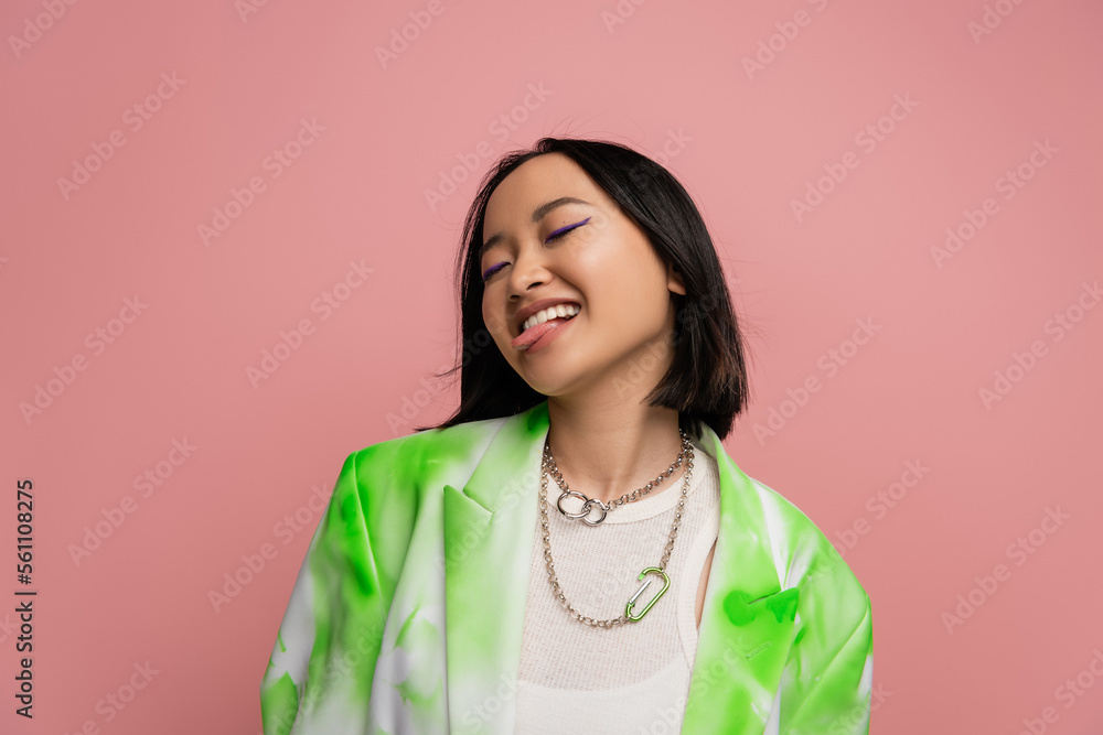 cheerful asian woman in green and white jacket and metal necklaces sticking out tongue isolated on pink. cheerful asian woman in green and white jacket and metal necklaces sticking out tongue isolated on pink.