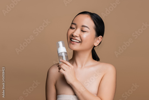 Positive asian model with naked shoulders holding cleansing foam isolated on brown.