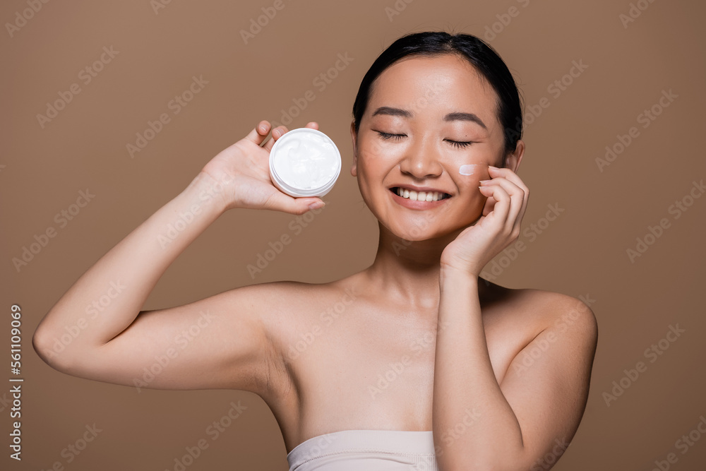 Positive asian woman with naked shoulders holding face cream isolated on brown. Positive asian woman with naked shoulders holding face cream isolated on brown.