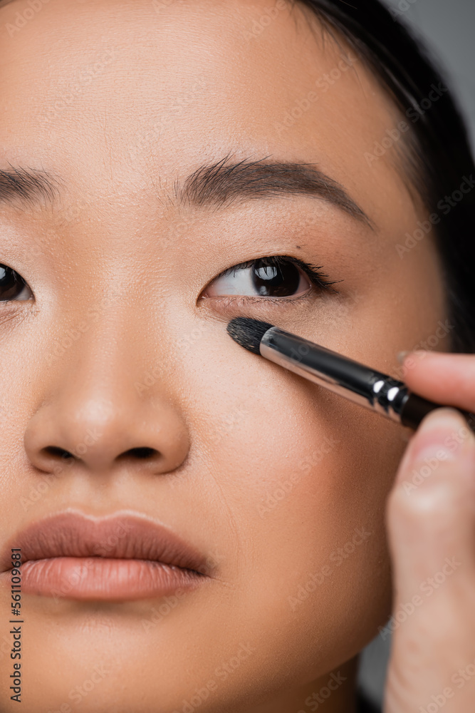 close up view of cropped asian woman with makeup foundation applying eye shadow isolated on grey.