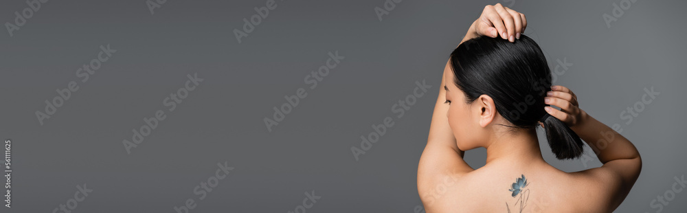 Naklejka premium Side view of young tattooed asian woman touching ponytail isolated on grey, banner.