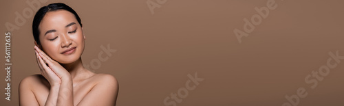 Pretty asian woman with naked shoulders holding hands near cheek isolated on brown, banner.
