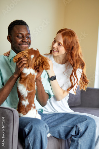 Cute young couple of black guy and caucasian lady sit on sofa having rest with pet dog. leisure time of nice friendly family. indoors. in domestic atmosphere. happy male and female with dog